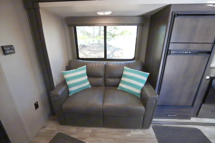 Inside an RV rental, a comfy brown sofa with teal pillows faces a large window, beside a dark wood refrigerator.. Grand Design Transcend Xplor 2020