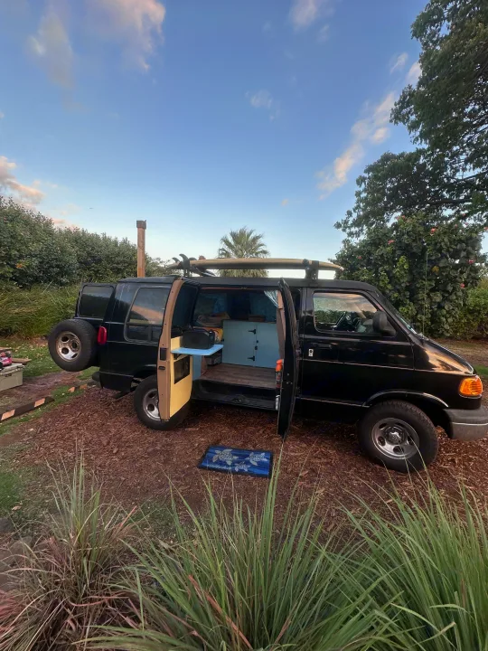 This black camper van, an RV rental, with surfboards on top, doors open revealing its custom interior. Parked for adventure.. Dodge 1500 1999