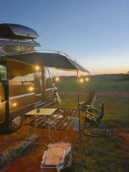Cozy RV rental setup at dusk: camper van with illuminated awning, table, chairs, and rug on grass under twilight sky.. Pleasure-Way Pleasure-Way Class B 2022