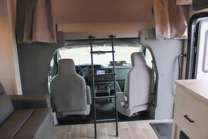 RV rental interior looking towards the cab: driver/passenger seats, dashboard, overhead bunk bed with ladder, sofa, and counter.. East to West Entrada 2023