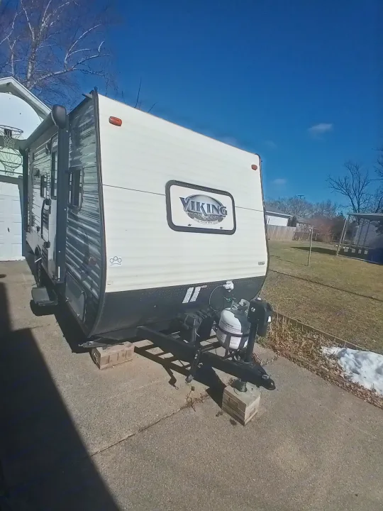 White & black Viking camper/RV rental, parked on a driveway. Features propane tanks & blocks. Clear blue sky overhead.. Coachmen Viking 2017