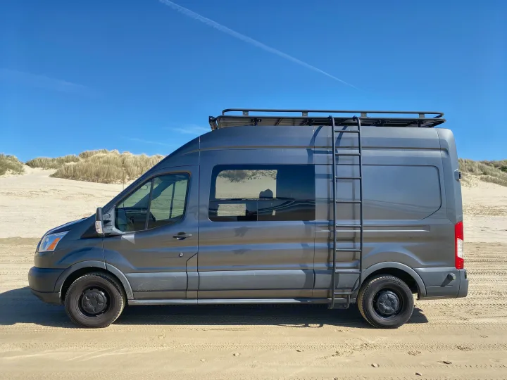 A dark gray camper van, perfect for an RV rental, parked on a sunny sandy beach with tall dunes under a clear blue sky.. Ford Transit 2016