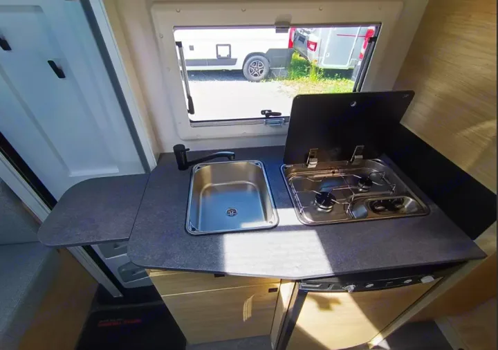 Compact RV rental kitchen: a sink, two-burner stove, and extendable counter, illuminated by a window view.. Challenger C194 START EDITION 2025
