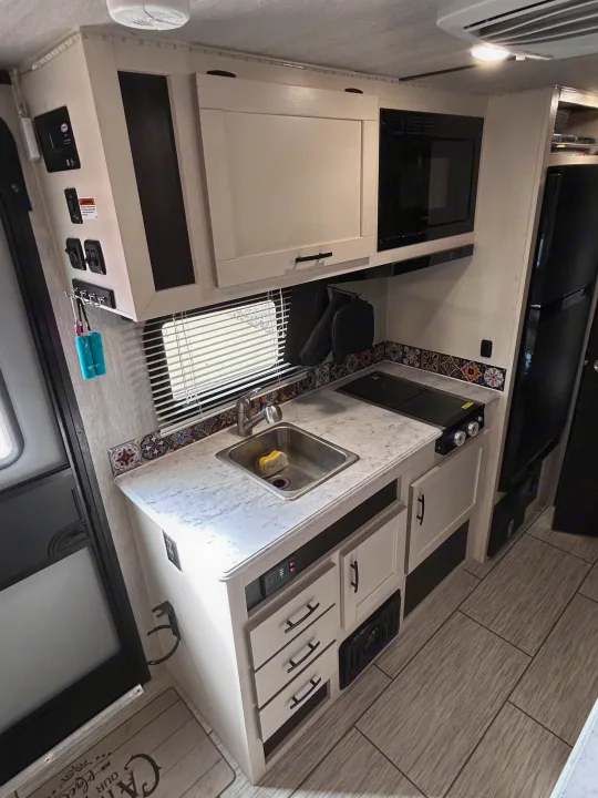 RV rental kitchen: light wood-look floor, light cabinets, stainless sink, microwave, and decorative tile backsplash.. Genesis Supreme 16 BB 2024