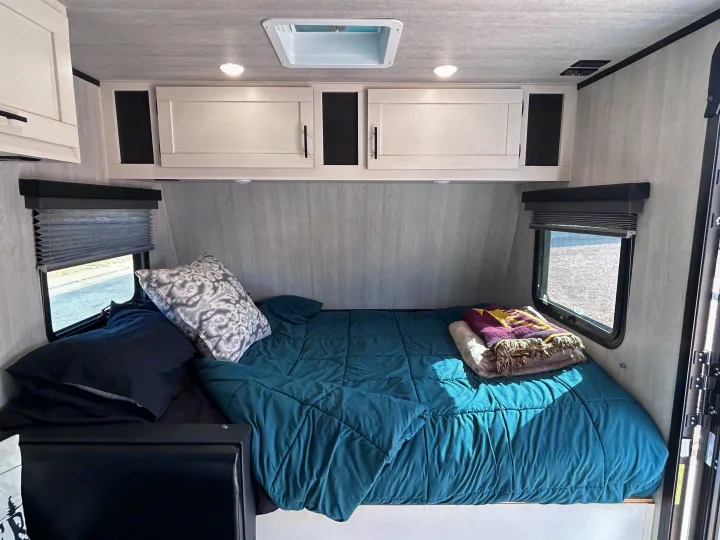 Inside a comfortable RV rental bedroom featuring a teal comforter, crisp white cabinets, and windows with blinds.. Genesis Supreme 16 BB 2024