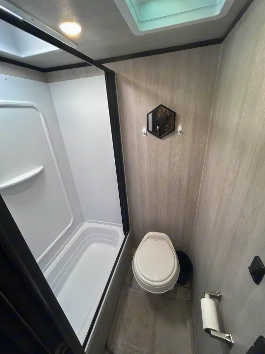 Compact RV rental bathroom featuring a white shower stall, toilet, and small hexagonal mirror on wood-grain walls.. Genesis Supreme 16 BB 2024