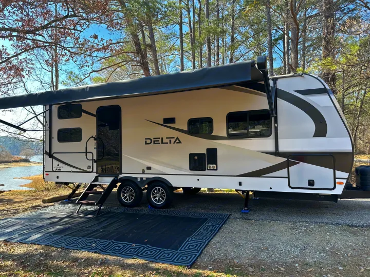 An Alliance Delta RV rental, neatly set up with an outdoor mat by a tranquil lake, awaits your camping adventure.. Alliance RV Delta 251BH 2024