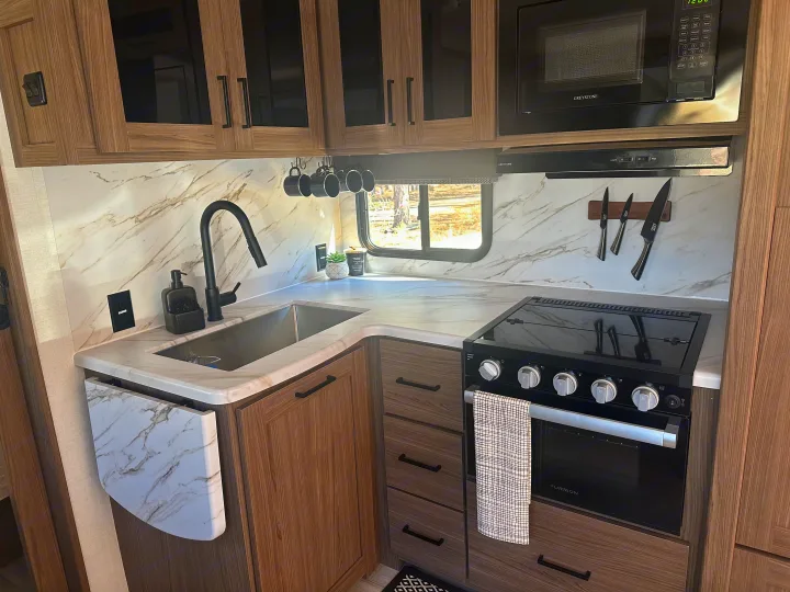 Modern kitchen in this RV rental, with marble-look counters, full appliances like stove, microwave, sink, and wood cabinets.. Alliance RV Delta 251BH 2024