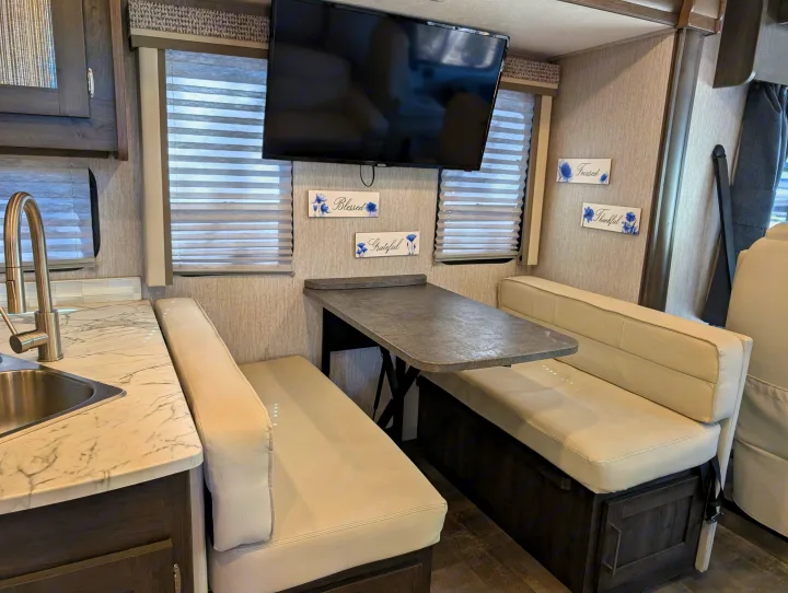 Bright and modern RV rental dining area with dual benches, a central table, wall-mounted TV, and adjacent kitchen.. Coachmen Pursuit 2022