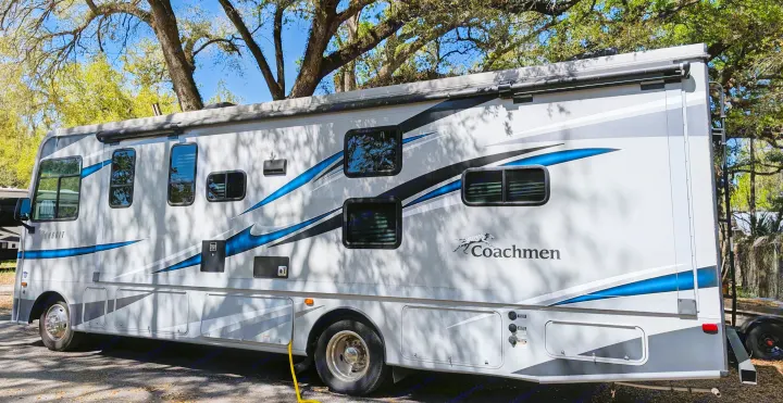 Side view of a modern white & gray Coachmen RV, perfect for your next rv rental camping trip, parked under trees.. Coachmen Pursuit 2022