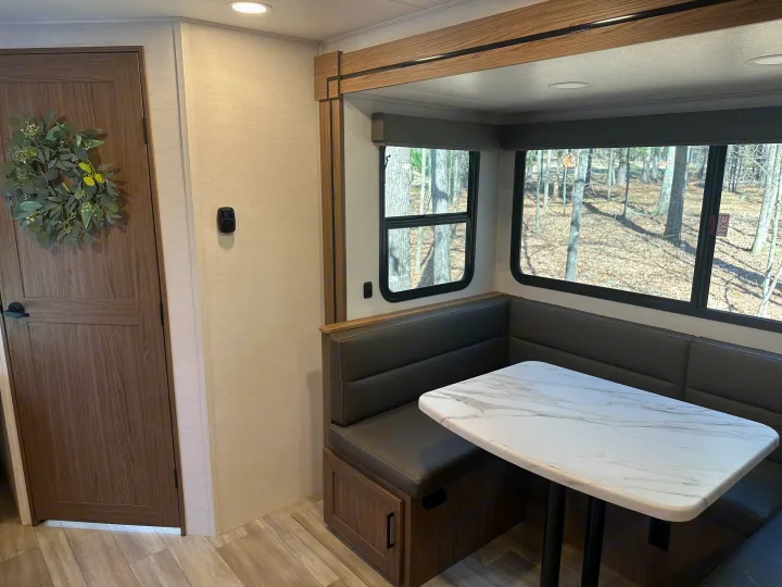 Cozy RV rental dinette with U-shaped seating, table, and large windows offering wooded views.. Alliance RV Delta 251BH 2024