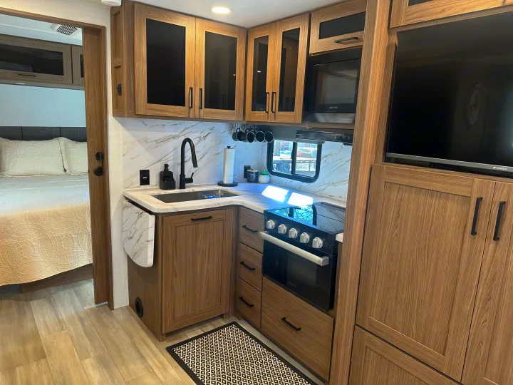 RV rental's modern kitchen features wood-grain cabinets, marble counters, black appliances, and a connected bedroom.. Alliance RV Delta 251BH 2024
