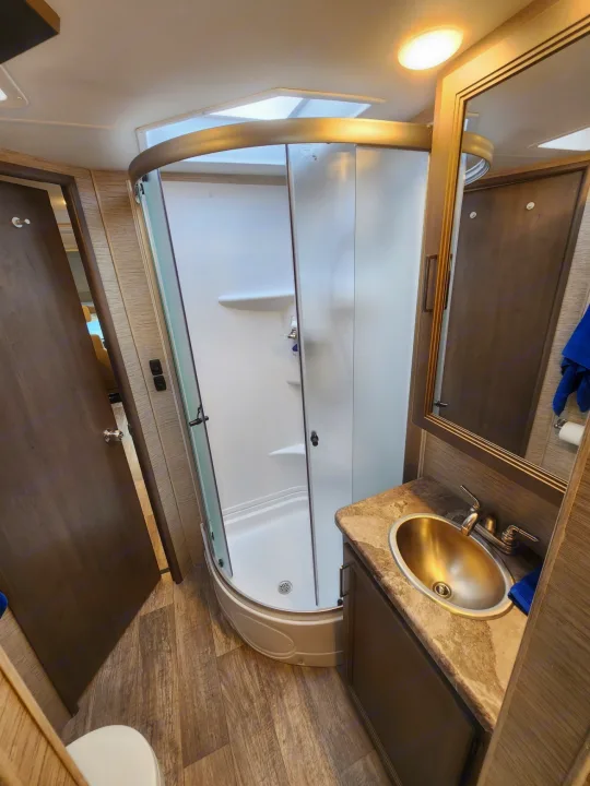 Compact RV rental bathroom with a corner glass shower, vanity sink, mirror, and toilet visible on wood-look floor.. Thor Motor Coach Chateau 2023