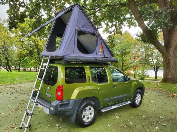A green Nissan Xterra with a deployed rooftop tent and ladder. Ideal RV rental for camping adventures by the water.. Nissan XTerra 2012