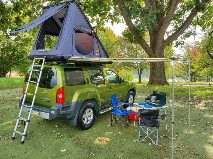 Green Xterra with a rooftop tent, extended awning, and camp setup including stove & chairs. An alternative to an RV rental for vehicle camping.. Nissan XTerra 2012