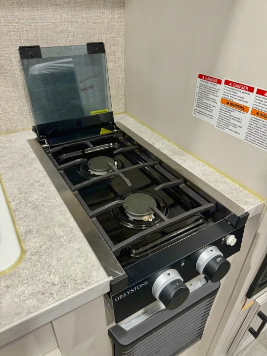 A Greystone two-burner gas cooktop with its glass lid open in an RV kitchen, suitable for an RV rental. Safety warnings are visible on the wall.. Jayco Jay Flight SLX 2022