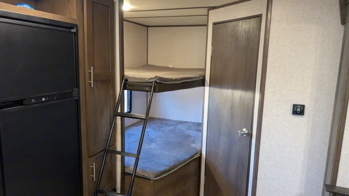 An RV rental interior showcasing practical bunk beds with ladder, integrated refrigerator, and warm wood cabinetry.. Dutchmen Coleman Lantern LT 2021