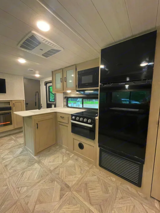 Explore this modern RV rental's spacious kitchen, featuring sleek black appliances, light wood-look cabinets, and unique flooring.. Forest River Wildwood X-Lite Platinum 2024
