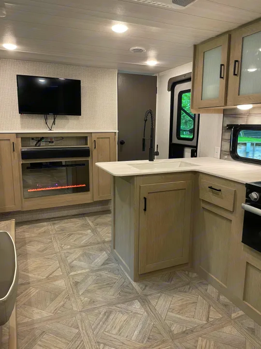 Modern RV rental interior: bright kitchen with island sink & black faucet, and living area with TV & fireplace.. Forest River Wildwood X-Lite Platinum 2024