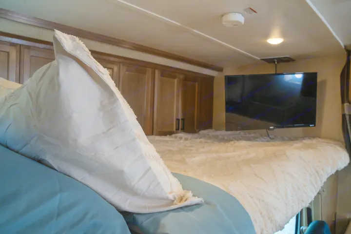 Comfortable cab-over bed in an RV rental, featuring a white comforter, blue sheets, mounted TV, and cabinets.. Gulf Stream Conquest 2019