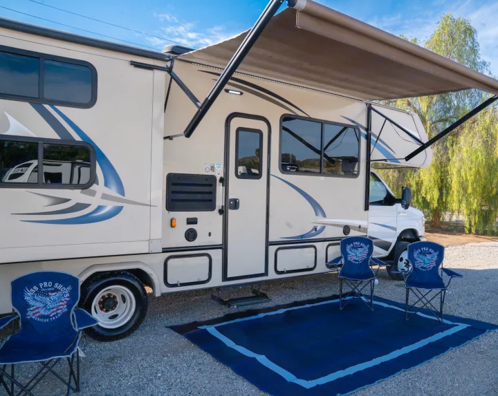 Enjoy an RV rental: Beige RV with extended awning, blue mat, and two Bass Pro Shops chairs on gravel.. Gulf Stream Conquest 2019