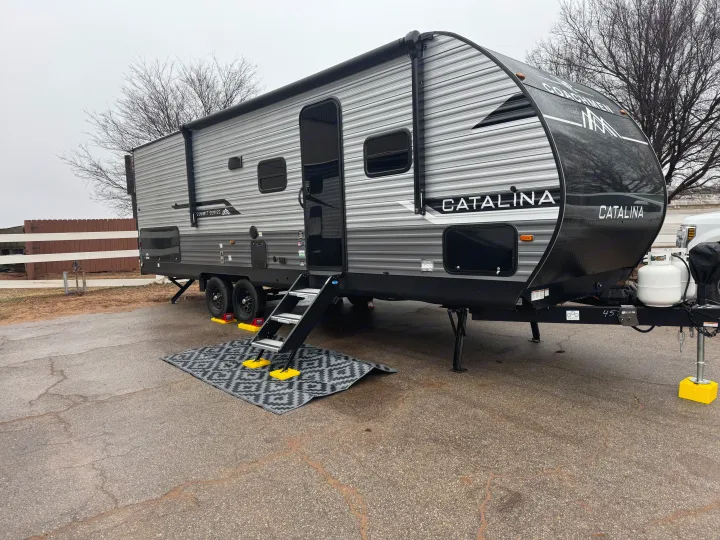 A gray Coachmen Catalina RV rental is parked on asphalt, featuring steps, an outdoor mat, and leveling gear.. Coachmen Catalina Summit Series 8 261BHS 2025