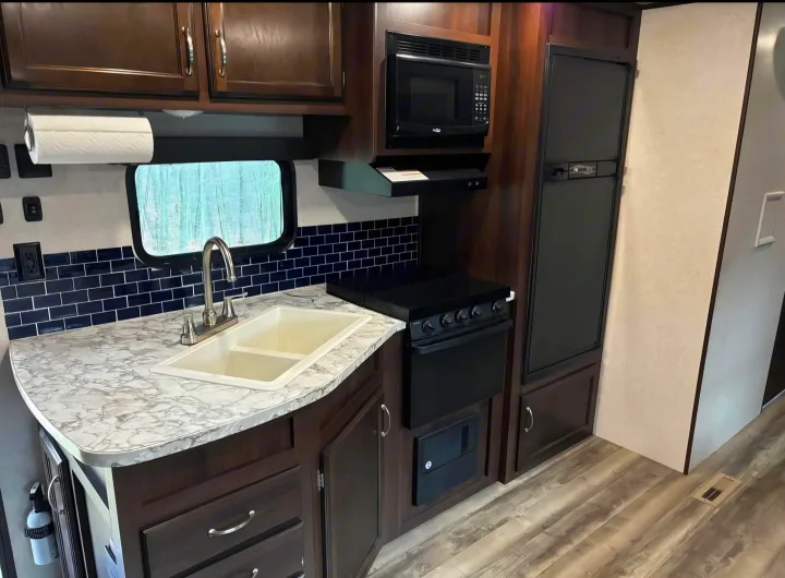 Modern RV rental kitchen featuring a sink, microwave, stove, fridge, dark wood cabinets, and blue tile backsplash.. Jayco Jay Flight SLX 2018