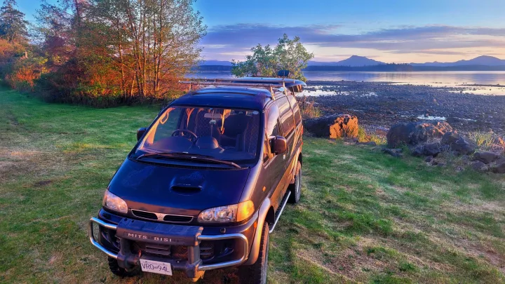 Mitsubishi van RV rental on grass by a scenic rocky bay with distant mountains at sunrise/sunset. Perfect for outdoor adventures.. MITSUBISHI Delica 1997