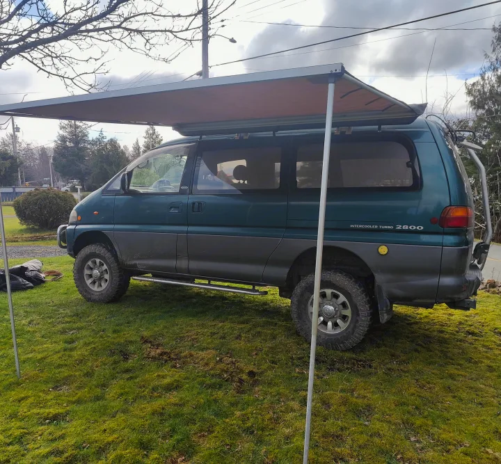 Rugged green Mitsubishi Delica 4x4 camper van with an extended awning set up on a grassy area. Perfect for an RV rental adventure.. MITSUBISHI Delica 1997
