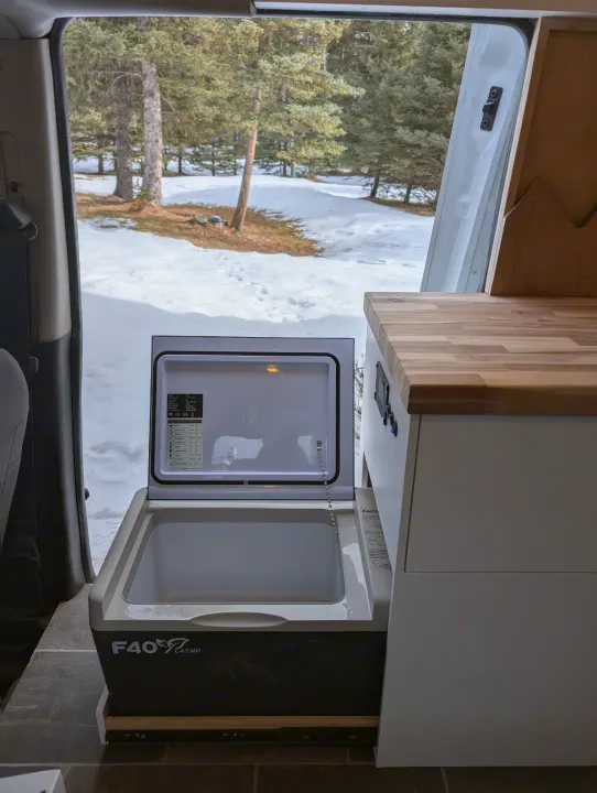 Inside an RV rental: a portable fridge open, set against a snowy forest. Perfect for mobile living adventures.. Dodge Sprinter Van 2015