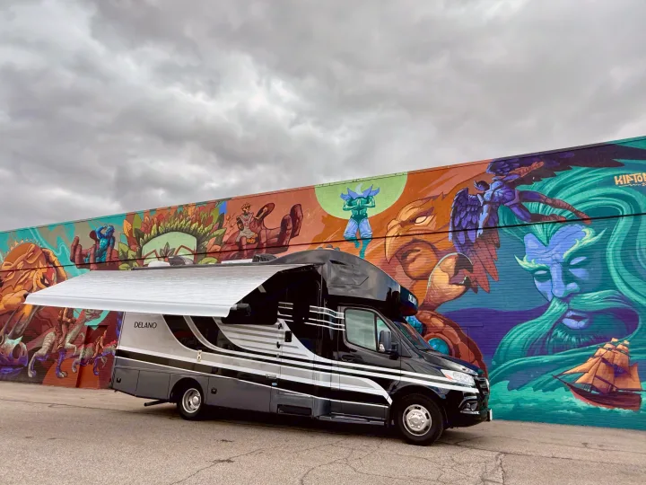 Stylish black and grey RV rental with extended awning, parked before a vibrant mural of mythical figures & a ship under cloudy skies.. Thor Delano 2023