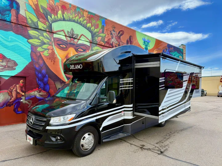 This sleek black Delano rv rental with an extended slide-out stands before a colorful brick mural.. Thor Delano 2023