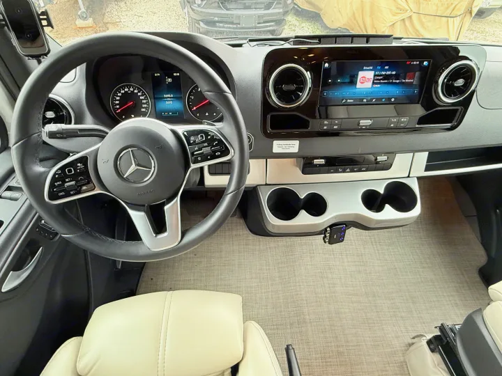 Modern rv rental cockpit view: Mercedes steering wheel, digital dash (5695 miles), and large infotainment screen with music playing.. Thor Delano 2023