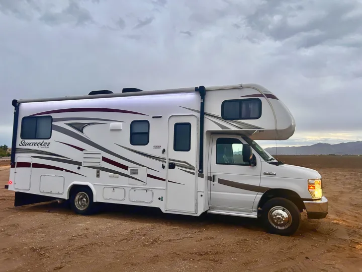 A white Sunseeker Class C motorhome, an ideal rv rental, sits on a dirt field with its lights on under a cloudy sky.. Forest River Sunseeker 2017