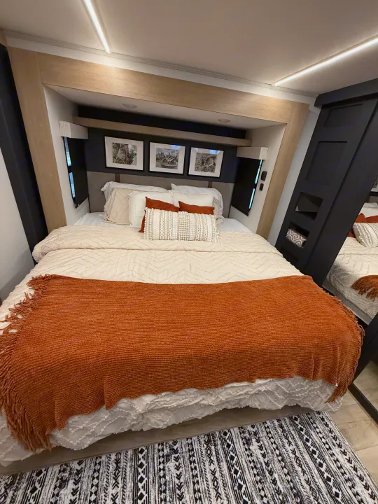 Relaxing RV rental bedroom showcasing a neutral bed, rust throw, framed art, and built-in storage.. Heartland Cyclone 2025
