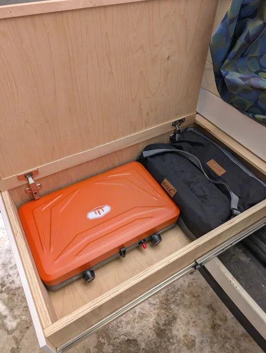 RV rental interior: A pull-out drawer reveals an orange GSI camping stove and black Nice C gear bags, neatly organized.. Dodge Sprinter Van 2015