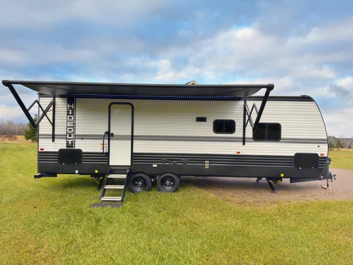 A sleek gray and black travel trailer with an extended awning, parked on green grass. Ideal for an RV rental adventure.. Keystone RV Hideout 2024