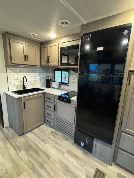 Modern RV rental kitchen with grey cabinets, subway tile, black sink, microwave, cooktop, and large refrigerator.. Keystone RV Hideout 2024