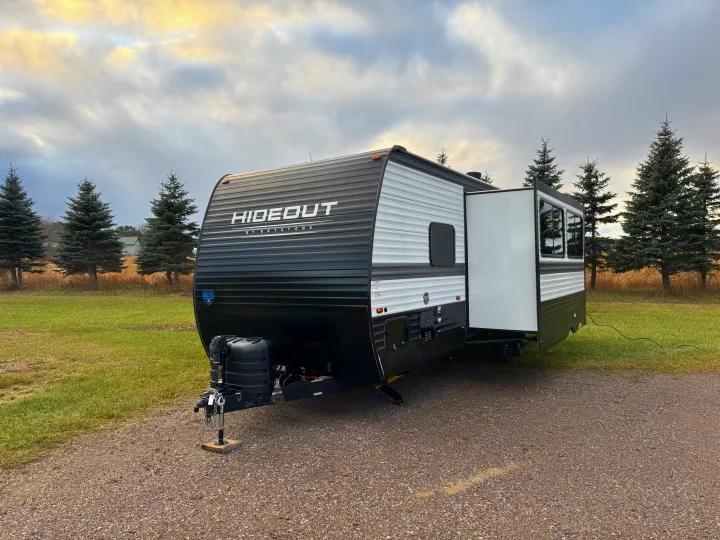 Black & white Hideout by Keystone RV rental, with an open slide-out, parked on gravel in a green field under cloudy skies.. Keystone RV Hideout 2024