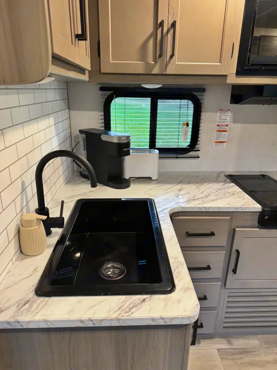 Stylish RV rental kitchen: black sink, Keurig, white subway tile backsplash, and light wood cabinets.. Keystone RV Hideout 2024
