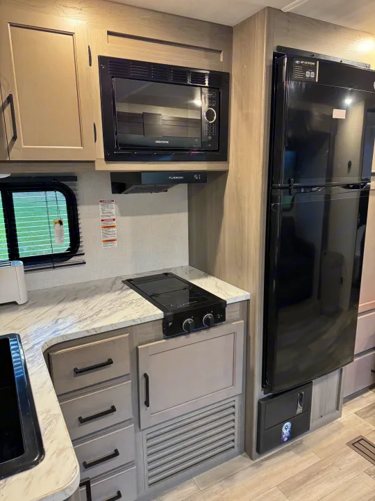 Modern kitchen setup in an RV rental, featuring a microwave, cooktop, black refrigerator, and light wood cabinetry.. Keystone RV Hideout 2024