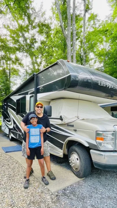 A dad and child smile in front of their large RV rental, parked under tall trees for a family camping adventure.. Forest River Coachmen Leprechaun 2020