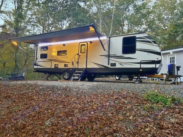 Illuminated rv rental travel trailer with awning extended, parked in a leafy, wooded campsite at dusk. Perfect for outdoor adventures.. Keystone RV Outback Ultra-Lite 2021