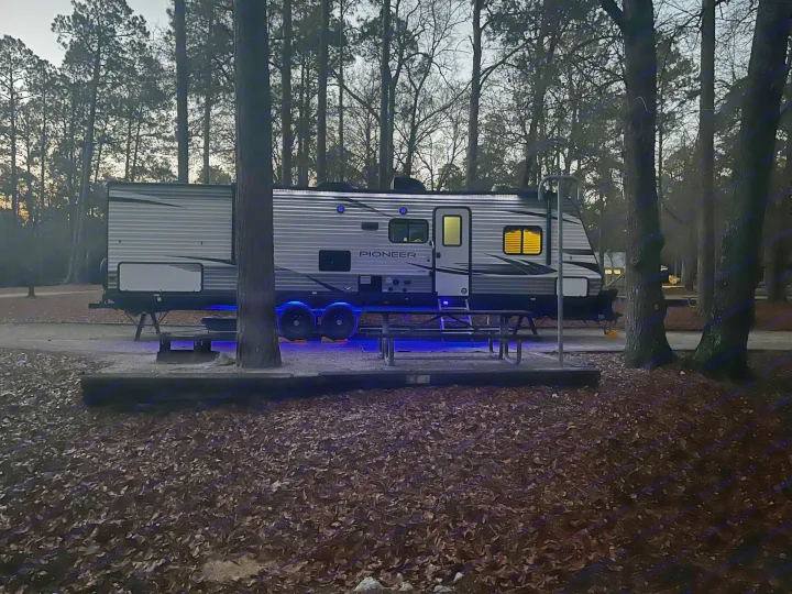 Experience nature with an RV rental. A modern travel trailer with blue and yellow lights brightens a serene, leafy campsite at dusk.. Heartland RVs Pioneer 2020