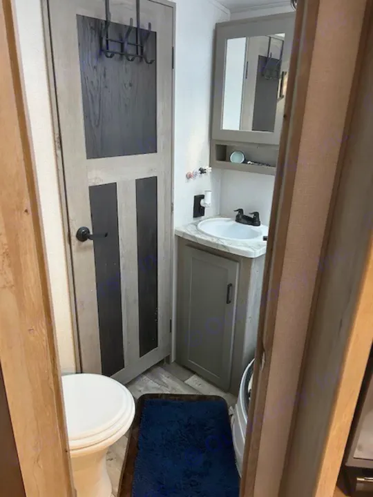 Peek into an RV rental bathroom: toilet, sink, mirror, and door with hooks. Dark blue mat on floor.. Keystone RV Outback Ultra-Lite 2021