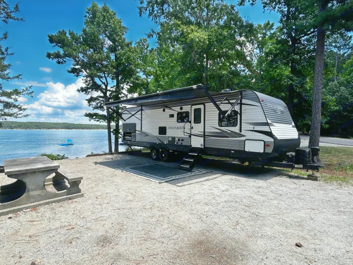 Lakeside RV rental getaway: a travel trailer at a gravel campsite with a picnic table, surrounded by trees and water.. Heartland RVs Pioneer 2020