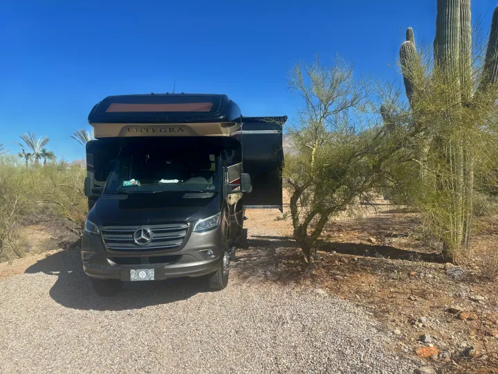 A dark Mercedes-Benz Sprinter rv rental motorhome is parked on gravel in a sunny desert landscape with cacti and trees.. Entegra Coach Qwest 2020