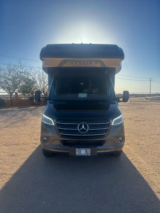 Front view of a dark brown Mercedes-Benz Entegra RV rental, parked on gravel with bright sun behind it, casting a long shadow.. Entegra Coach Qwest 2020