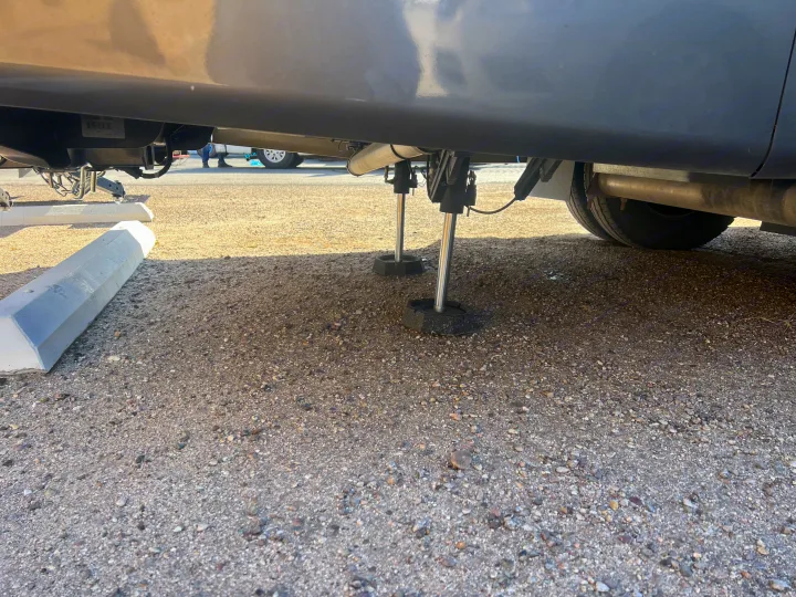 RV rental with hydraulic leveling jacks deployed on a gravel lot, viewed from a low angle under the vehicle.. Entegra Coach Qwest 2020