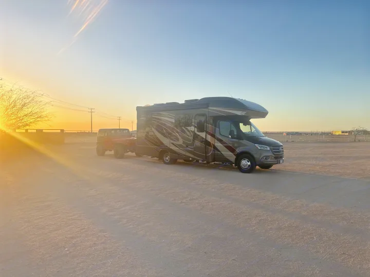 An RV rental tows a red Jeep, parked in a vast dirt lot during a beautiful golden sunset.. Entegra Coach Qwest 2020